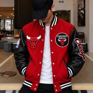 May include: A red and black varsity jacket featuring the Chicago Bulls logo on the chest and sleeve, an NBA patch on the right shoulder, and white striped ribbed trim on the collar, cuffs, and hem, worn with a plain white t-shirt and black cap.