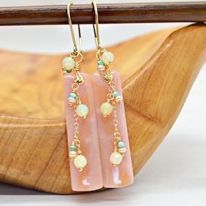 May include: A pair of long, rectangular earrings with a pale pink hue. The earrings feature gold-colored hooks and chains, adorned with small, round, opalescent beads, turquoise beads, and tiny gold accents. The earrings are displayed on a wooden surface.