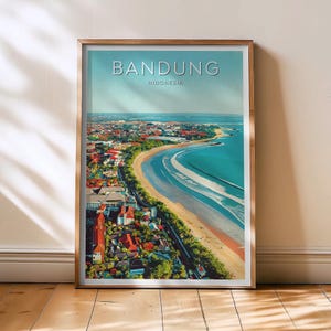 Op de afbeelding: Ingelijste print van Bandung, Indonesië, met een kustzicht met een zandstrand en turquoise water. De stad is zichtbaar met gebouwen en rode daken. De print heeft een lichtblauwe lucht en het woord "BANDUNG" staat bovenaan.