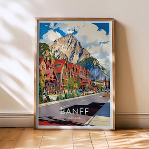 Può includere: Una stampa incorniciata con una vista panoramica di Banff, Canada. L'opera raffigura una strada fiancheggiata da edifici con tetti rossi, uno sfondo montuoso e il testo "BANFF CANADA". La stampa è in una cornice dorata.