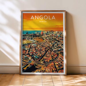 May include: A framed print featuring a vibrant aerial view of Angola, Africa. The artwork showcases a cityscape with colorful buildings, a coastal horizon, and the text "ANGOLA AFRICA" in white. The print is displayed on a wooden floor.