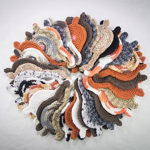 May include: A collection of crocheted cat-shaped ornaments in various shades of brown, orange, white, and gray. The ornaments are arranged in a circle, creating a decorative pattern.