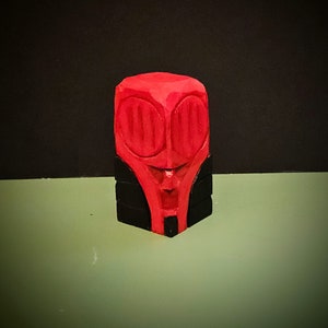 May include: A red wooden sculpture with a face carved into it. The sculpture has black accents around the eyes and mouth.