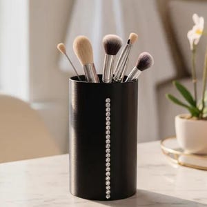 Puede incluir: Un soporte cilíndrico negro para brochas de maquillaje con una línea vertical de pequeños diamantes de imitación transparentes. El soporte contiene varios pinceles de maquillaje con varios cabezales. El soporte está sobre una superficie de mármol blanco.