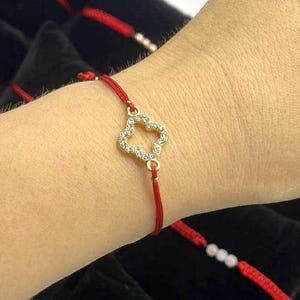Puede incluir: Una pulsera de hilo rojo con un dije de trébol dorado, adornada con pequeños cristales transparentes. Otras pulseras de hilo rojo con dijes dorados son visibles en el fondo.