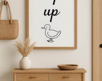 May include: A framed print with the words "ey up" and a duck illustration. The print hangs above a wooden console table with two drawers and a lower shelf. A woven basket bag hangs on the wall to the left.