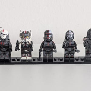 Könnte beinhalten: Fünf Kunststoff-Klemmbaustein-Figuren, die gepanzerte Soldaten darstellen. Jede Figur trägt einen einzigartigen Helm und taktische Ausrüstung in Schwarz, Weiß und Rot. Sie stehen auf einzelnen schwarzen, rechteckigen Sockeln und halten kleine Spielzeugwaffen.