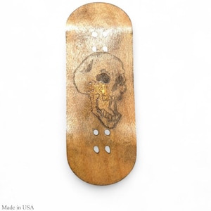 May include: A wooden fingerboard with a skull design. The board is a light brown color with a skull etched into the wood. The skull is a detailed drawing with dark lines. The fingerboard has four holes for the trucks. The text "Made in USA" is at the bottom.
