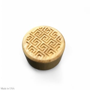 May include: A round, light brown bottle stopper with a carved geometric pattern on the top surface. The stopper is cylindrical with a flat base and a detailed design. The text "Made in USA" is visible at the bottom left.