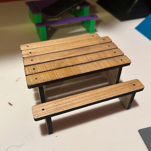 May include: A miniature wooden picnic table with a light brown tabletop and bench, supported by black legs. A small black skateboard rests on a purple and green miniature picnic table in the background. The scene suggests a playful, miniature world.