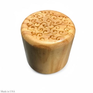 May include: A cylindrical wooden bottle stopper with a carved swirling pattern on the top. The stopper is made of light-colored wood and has a smooth, polished finish. The text "Made in USA" is visible at the bottom.