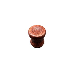 May include: A close-up of a wooden ear plug. The plug is a dark reddish-brown color with visible wood grain. The shape is a flared cylinder, wider at the ends and narrower in the middle. The background is white.