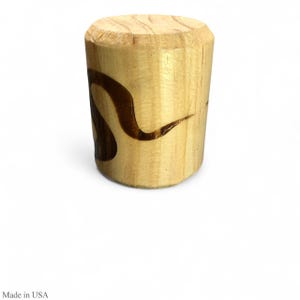 Könnte beinhalten: Ein zylindrischer Weinflaschenverschluss aus Holz mit einem dunkelbraunen, stilisierten Design. Der Verschluss ist aus hellfarbenem Holz gefertigt und hat eine glatte, abgerundete Oberseite. Das Design scheint in das Holz eingebrannt zu sein.