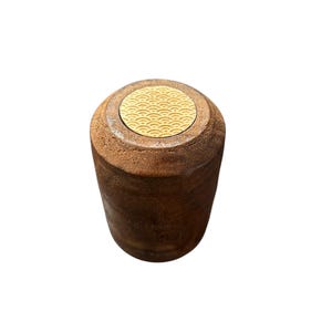 May include: A cylindrical, brown wooden container with a circular, gold-colored top. The top features a repeating wave pattern. The container has a rustic, handcrafted appearance, suggesting it may be used for storage or display.