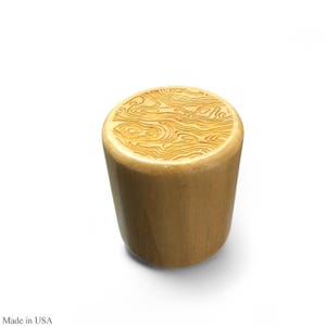 May include: A cylindrical wooden bottle stopper with a topographic map design etched on the top. The stopper is made of light-colored wood and has a smooth, rounded shape. The image is set against a white background.