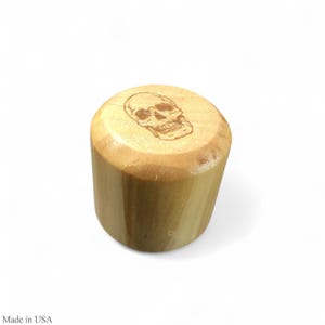 May include: A cylindrical wooden bottle stopper with a skull design etched on the top. The stopper is made of light-colored wood and has a natural finish. The skull is a detailed illustration.