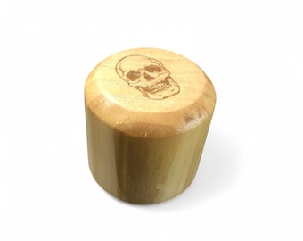 May include: A cylindrical wooden bottle stopper with a skull design etched on the top. The stopper is made of light-colored wood and has a natural finish. The skull is a detailed illustration.