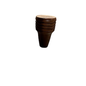 Puede incluir: Un tapón de botella de madera marrón oscuro con una parte superior redondeada y un cuerpo cilíndrico acanalado. El tapón se estrecha hasta una forma cónica en la parte inferior. La veta de la madera es visible en la parte superior.