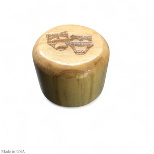 May include: A light-colored wooden bottle stopper with a theatrical mask design etched on the top. The stopper is cylindrical with a slightly rounded top and a smooth, natural finish. The design features two stylized masks, one smiling and one frowning.
