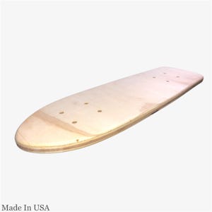 Könnte beinhalten: Ein Skateboard-Deck aus Naturholz mit abgerundeter Nase und Heck. Das Deck hat vorgebohrte Löcher für Trucks und wird in den USA hergestellt. Die Holzmaserung ist sichtbar und das Deck scheint unfertig zu sein.