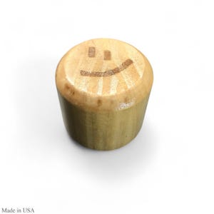 May include: A cylindrical wooden bottle stopper with a smiling face etched on the top. The stopper is made of light-colored wood and has a smooth, rounded surface. The text "Made in USA" is visible at the bottom.
