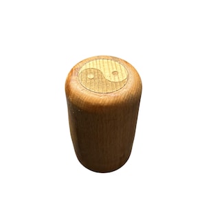 May include: A cylindrical wooden bottle stopper with a carved Yin and Yang symbol on the top. The stopper is made of light brown wood and has a smooth, polished finish. The Yin and Yang symbol is a lighter shade of brown.