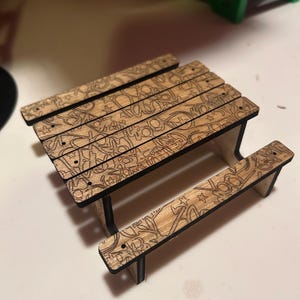 May include: A miniature wooden picnic table with engraved designs. The table has a light brown surface with black edges and legs. The picnic table has a bench on each side and is suitable for small figurines.