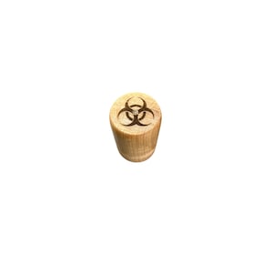 May include: A cylindrical wooden bottle stopper with a biohazard symbol etched on the top. The stopper is made of light-colored wood and has a smooth, rounded shape. The biohazard symbol is a dark brown color.
