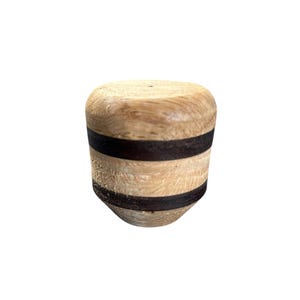 May include: A wooden bottle stopper with a light brown top and bottom, and two dark brown bands. The stopper has a cylindrical shape with a slightly tapered base. The wood grain is visible.