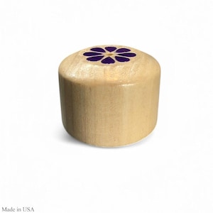 May include: A cylindrical wooden bottle stopper with a purple flower design on top. The stopper is made of light-colored wood and is suitable for wine bottles. The text "Made in USA" is printed at the bottom.
