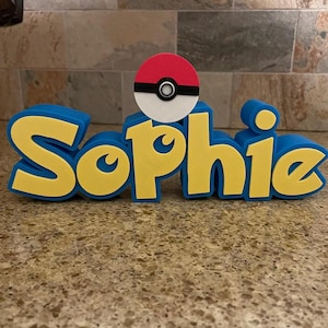 May include: A blue and yellow 3D name sign that reads "Sophie". A red, white, and black Pokeball sits atop the name. The sign is a decorative item.