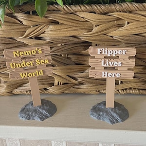 May include: Two decorative wooden signs with the text "Nemo's Under Sea World" and "Flipper Lives Here". The signs are brown with gold lettering and are mounted on gray rock bases. A woven basket and greenery are in the background.