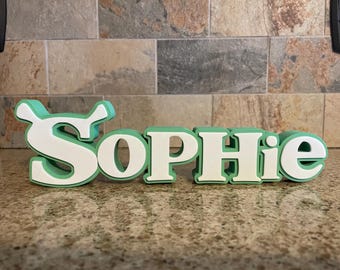 May include: A 3D-printed nameplate that spells out "Sophie" in white letters with a green outline. The nameplate is displayed on a speckled countertop with a brick-patterned wall in the background.