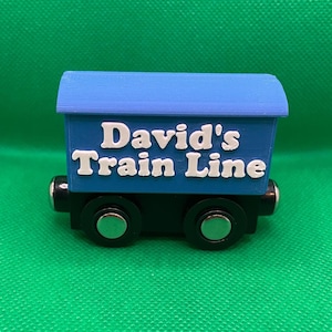 May include: A blue toy train car with the text "David's Train Line" in white. The train car has a blue roof, a dark blue body, and black wheels with silver accents. The toy is designed for children.