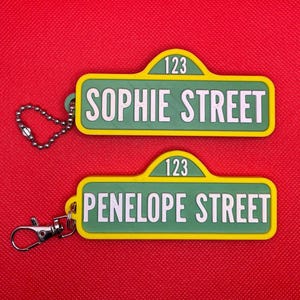 May include: Two keychains resembling street signs, one reading "SOPHIE STREET" and the other "PENELOPE STREET." Both have a green background with white text and a yellow border. Each keychain has a silver chain or clasp.