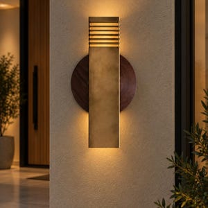May include: A rectangular wall sconce featuring a brushed brass finish with a slatted top section. The fixture is mounted on a circular dark wood backplate and emits a warm, soft glow from both the top and bottom edges of the metal body.