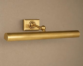 May include: A brass picture light fixture featuring a rectangular wall-mounted base, a single adjustable arm with a ball joint, and a long, cylindrical horizontal shade with a smooth, metallic finish and a warm, golden-toned appearance.