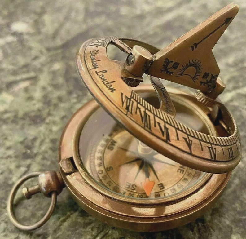May include: A vintage brass compass with a sundial feature. The compass has a hinged lid with Roman numerals and the text "Stanley London". The sundial arm is etched with a sun and arrow design. The compass face shows directional markings.
