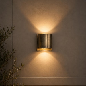 May include: A cylindrical wall sconce with a brushed brass finish mounted on a flat surface. The fixture emits a warm, glowing light that projects upwards and downwards in two distinct beams, creating a symmetrical pattern of illumination.
