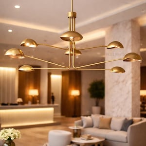 Puede incluir: Una lámpara de araña moderna, dorada, con múltiples brazos curvos, cada uno con una lámpara en forma de cúpula. La lámpara de araña cuelga en un interior contemporáneo y bien iluminado. La imagen también muestra una zona de estar con sofás y mesas.