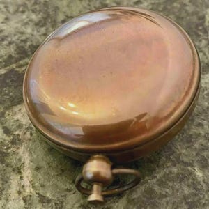 May include: A round, antique-style compass with a copper-colored finish. The compass has a domed top and a small ring attached to the base. The metal has a polished, reflective surface.