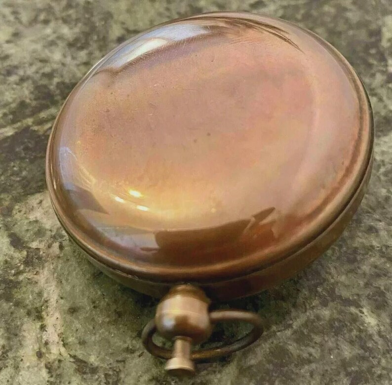 May include: A round, antique-style compass with a copper-colored finish. The compass has a domed top and a small ring attached to the base. The metal has a polished, reflective surface.