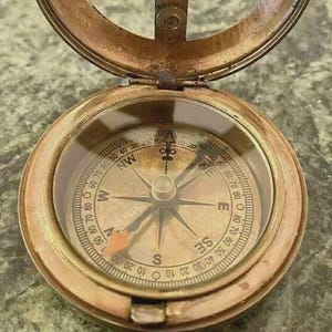 May include: Antique brass compass with a hinged lid, featuring a detailed compass rose and directional markings. The compass has a clear glass cover and a small ring for attachment. The compass is a navigational tool.