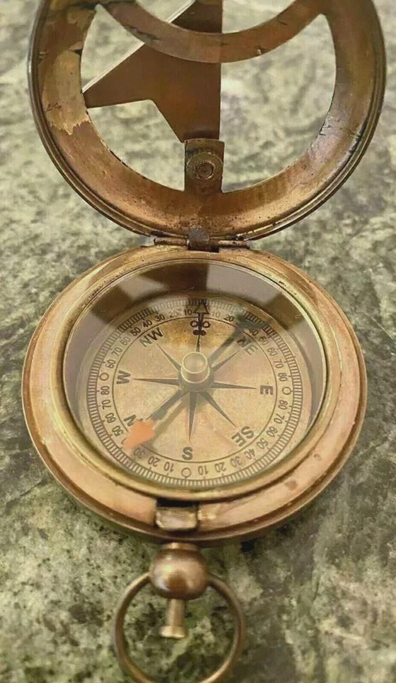 May include: Antique brass compass with a hinged lid, featuring a detailed compass rose and directional markings. The compass has a clear glass cover and a small ring for attachment. The compass is a navigational tool.