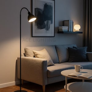 Op de afbeelding: Een zwarte vloerlamp met een gebogen arm en een ronde voet verlicht een woonkamer. De lamp staat naast een grijze bank met kussens. De tekst "ACE LIGHTING" is zichtbaar op de muur.