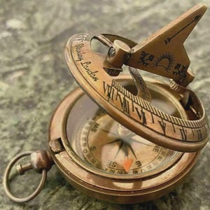 May include: A vintage brass sundial compass with a hinged gnomon and a compass dial. The sundial features Roman numerals and the inscription "Stanley London." The compass needle points north. The sundial is set against a green marble surface.