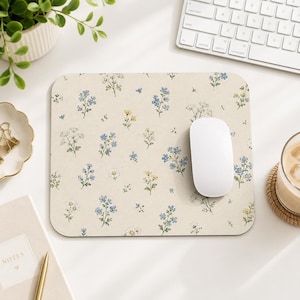 Op de afbeelding: Een rechthoekige muismat met een crèmekleurige achtergrond en een herhalend patroon van kleine blauwe, gele en witte wilde bloemen met groene stengels en bladeren. Een gladde witte computermuis rust aan de rechterkant van de mat.