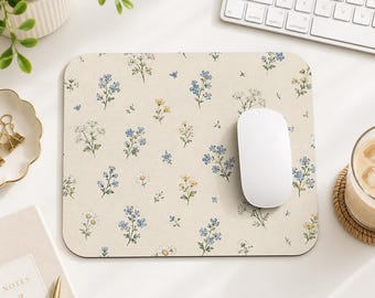 Puede incluir: Una alfombrilla de ratón rectangular con fondo color crema que presenta un patrón repetitivo de pequeñas flores silvestres azules, amarillas y blancas con tallos y hojas verdes. Un ratón de ordenador blanco liso descansa a la derecha.