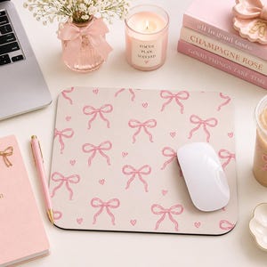 May include: A square mouse pad featuring a repeating pattern of light pink bows and small hearts on a pale background. The mouse pad is positioned on a desk surface alongside a white computer mouse, a pink notebook, a pen, and other desk accessories.