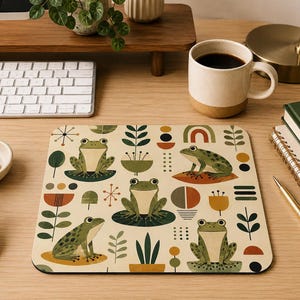 May include: A square mouse pad featuring a repeating pattern of green frogs on lily pads, stylized plants, and geometric shapes in shades of olive, mustard yellow, and burnt orange on a light cream background with rounded corners.